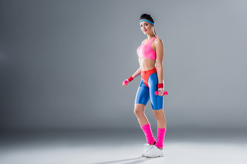 beautiful sporty girl holding dumbbells and smiling at camera on grey