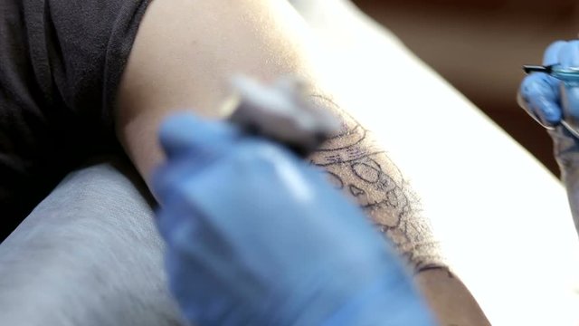 Close-up of a man with blue gloves doing a tattoo on his arm for a brutal man. Close-up of a needle with ink in the hand of a professional tattoo artist. Professional artist making tattoo in salon.