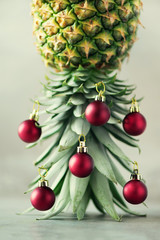 Obraz na płótnie Canvas Creative Christmas tree made of pineapple and red bauble on grey concrete background, copy space. Greeting card, decoration for new year party. Holiday concept.