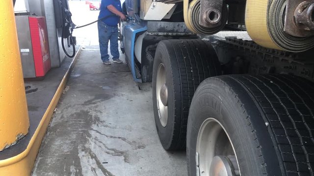 Truck driver pumps diesel fuel into semi at a filling station during daytime.  