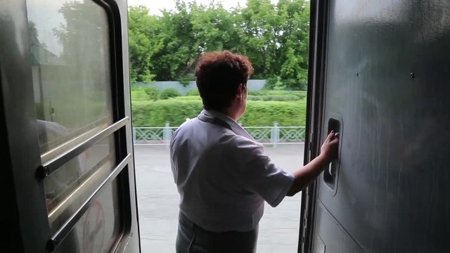 Train conductor stands at the door of the car. A female railway worker stands in the open doorway of the car while the train is moving.