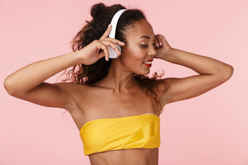 Woman in swimwear posing isolated over pink background listening music with headphones.