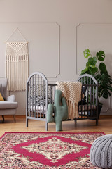 Patterned carpet in front of grey cradle with cactus pillow in baby's bedroom interior. Real photo