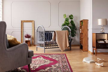 Armchair on carpet in grey kid's bedroom interior with blanket on cradle and lamp on cabinet. Real...