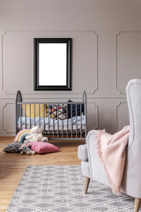 Mockup of empty poster above cradle in grey baby's bedroom interior with armchair. Real photo