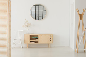 Round mirror above wooden cabinet with books and glass vase with flower, white chair next to it,...