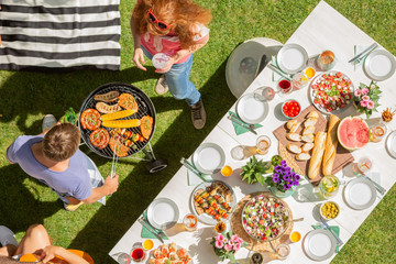 High angle on man and woman grilling shashliks next to table with food in the garden