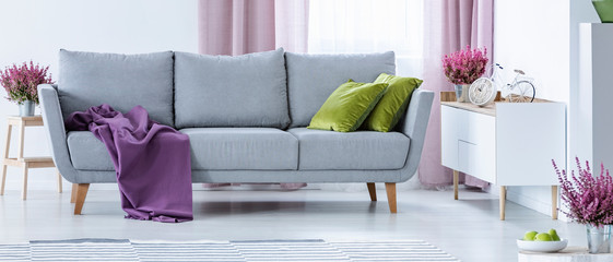 Grey couch with purple blanket and green pillows in real photo of white sitting room interior with fresh heathers and cupboard with bike shape clock