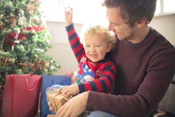Cute child sitting on his father leg, opening a christmas gift