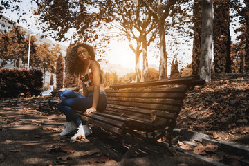 A charming curly woman hipster in glasses is sitting on the park bench, sunny autumn day; a...