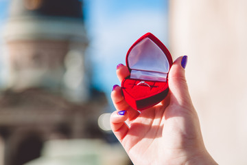 Womans hand is holding engagement ring with diamond in the red box of heart form in front of...