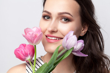 Portrait of young brunette beautiful woman with makeup in striped dress, standing and holding tulips and looking at camera with toothy smile. indoor studio shot, isolated on light grey background.