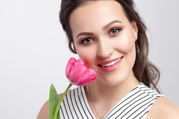 Portrait of young brunette beautiful woman with makeup in striped dress, standing and holding pink tulip and looking at camera with toothy smile. indoor studio shot, isolated on light grey background.