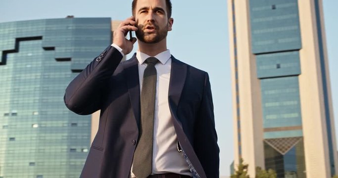 Multiethnic businessman walking down street between scyscrapers with briefcase in hand, talking on phone, and smiling closeup 4k