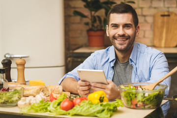 Man preparing delicious and healthy food in the home kitchen on a sunny day. Using tablet computer for searching recipes.