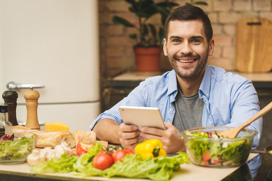 Man Preparing Delicious And Healthy Food In The Home Kitchen On A Sunny Day. Using Tablet Computer For Searching Recipes.