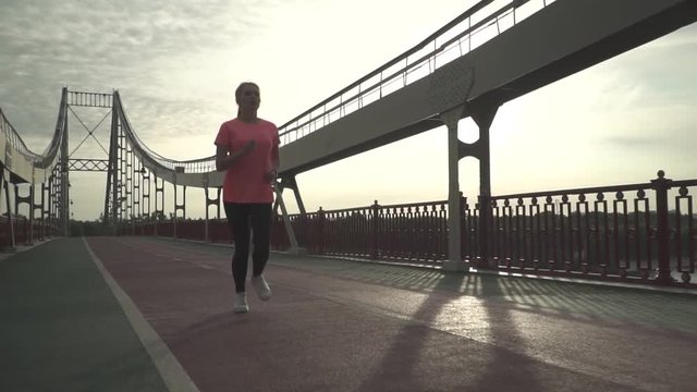 Athletic blonde girl is running on the bridge in the morning. Slow motion