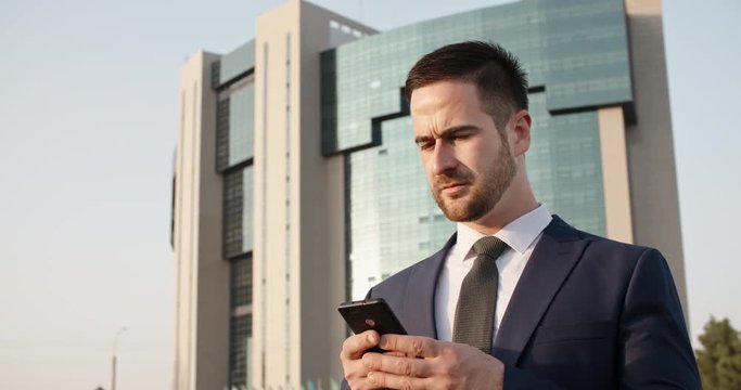 Confident multiethnic in suit and tie businessman texting on his smartphone via internet in the street - closeup 4k