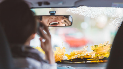 Young woman smiling and laughing to rear-view mirror in a car while talking on phone and driving. Shot from behind - Autumn mood - multitasking concept