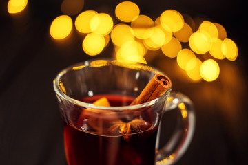 closeup piece of glass of mulled wine with orange and cinnamon on dark black background, on white plate, Christmas lights, large yellow bokeh