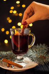 closeup glass of mulled wine with orange and cinnamon on dark black background, on white plate, christmas ball, tree and lights, cinnamon in female hand