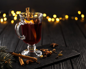 closeup glass of mulled wine with orange and cinnamon on dark black background,  Christmas tree and lights, large yellow bokeh, mulled wine set