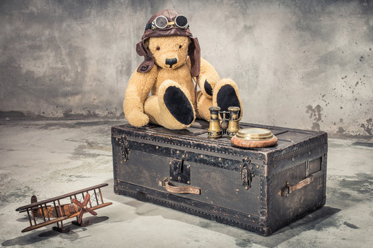 Teddy Bear with leather aviator's hat and goggles sitting on vintage old classic travel trunk circa 1900s with binoculars, compass, wooden toy plane. Travel by air concept. Retro style filtered photo