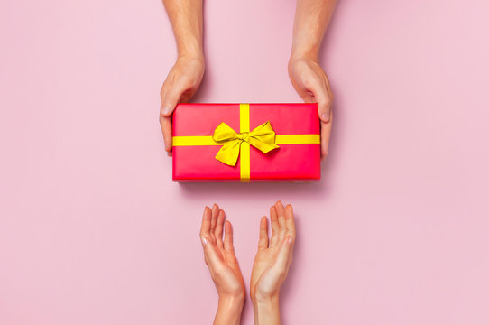 Top View Of Male And Female Hands Holding Red Gift Box With Golden Ribbon On Pink Background Flat Lay. Present For Birthday, Valentine Day, Christmas, New Year. Congratulations Background Copy Space.