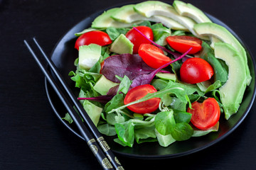 Vegetable salad with avocado, cherry tomatoes, arugula and spinach on a black plate on a dark background.