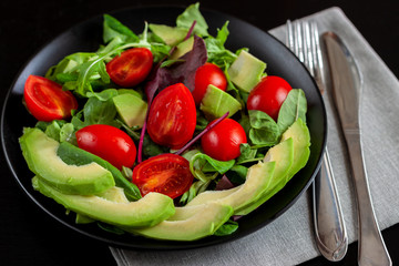 Vegetable salad with avocado, cherry tomatoes, arugula and spinach on a black plate on a dark background.