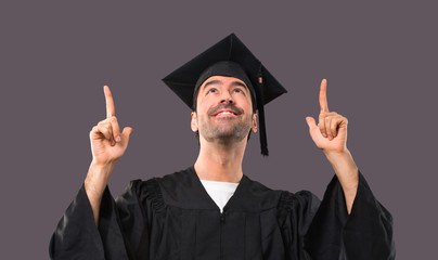 Man on his graduation day University pointing with the index finger a great idea and looking up on violet background