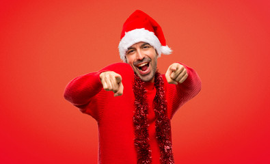 Man with red clothes celebrating the Christmas holidays points finger at you while smiling on red background