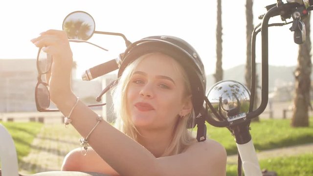 Young caucasian girl wearing helmet and protective glasses sits near electric bicycle on palm alley, blue sky and wide walkway.
