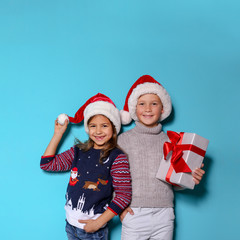 Cute children in warm sweaters and Christmas hats and gift on color background