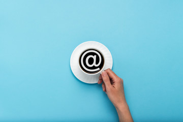 Female hand holding a white cup with black coffee on a blue background. Email sign. Flat lay, top view