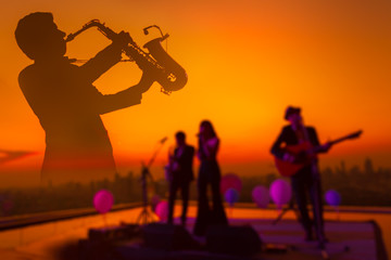 Fototapeta premium Fading silhouette saxophone musician man showing with blurry jazz trio band and twilight cityscape background. Double exposure process and copy space