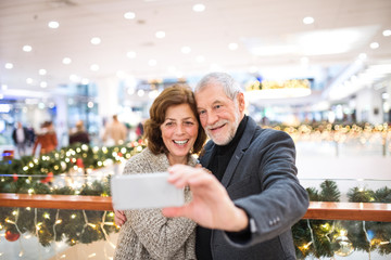 Senior couple with smartphone taking selfie in shopping center at Christmas time.