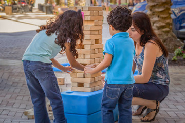The big sister removes the block of wood from the jumbling tower while little brother and mom watch.