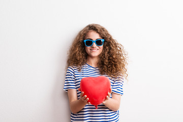 Young beautiful cheerful woman with sunglasses in studio, holding red heart.