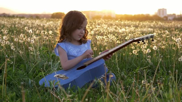 cute little girl sits in the grass and plays the jeans guitar. in the dandelions field spring bloom at sunset. concept music in nature away from the city