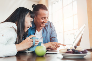 Happy couple sitting together at the table with laptop watching movies, with cookies, fruits and hot drinks. Multiethnic young family