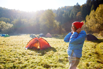 happy smiling woman in a blue down jacket is putting on a red cap. Woman is standing in campsite on a meadow in the autumn forest on a bright sunny foggy morning
