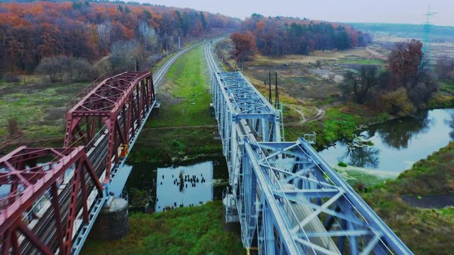 Aerial following shot of a suburban train passes the steel bridge over the river and enters the autumn forest. Back view