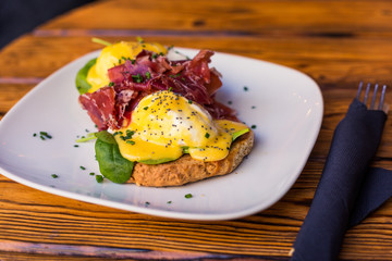 Two delicious poached eggs served with fried bacon for breakfast on a white plate, with spinach and onions. The dish is on a wooden table. Restaurant serving