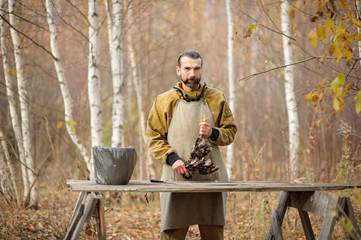 bearded white man in apron holding a slaughter chicken in his hands and  looking at the camera against the forest.
