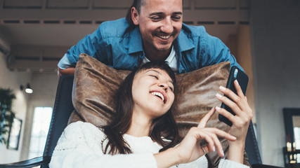 Obraz premium Couple sitting together in leather couch and taking selfie photos using mobile phone camera. Multiethnic young family