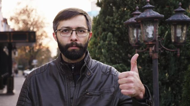 Portrait of young attractive bearded man in glasses and looking at the camera. Man with poker face making thumbs up