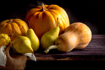 Large yellow pumpkins and pears lie on a rough cloth and gauze on a black wooden table and black background
