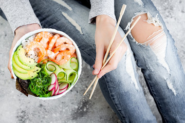 Girl in jeans holding shrimp poke bowl with seaweed, avocado, cucumber, radish, sesame seeds.