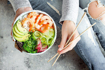 Girl in jeans holding shrimp poke bowl with seaweed, avocado, cucumber, radish, sesame seeds.
